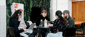 Five people sit around a café table with laptops, working together in a bright room with plants and chandeliers.