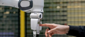 Human hand touches white robotic arm’s gripper in an indoor industrial setting