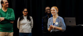 Smiling woman in blue striped shirt presenting beside three colleagues and an open laptop on stage
