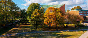 Campus park with green and orange autumn trees before a brick building under a blue sky with clouds