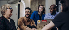 Five adults of varied ages sit indoors, smiling and talking around an open laptop on a wooden table.