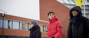 Three students in winter coats walk in snow by a red brick campus building with a yellow flag.