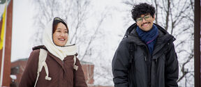 Two smiling students in winter coats walking outside on a snowy campus with bare trees and brick buildings.