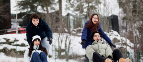 Four friends in winter coats sledding downhill on snow, laughing in front of a forest and cabins