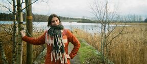 Person in orange coat and large scarf posing on lakeside path between bare trees and reeds