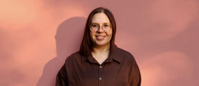 Smiling person in glasses and brown shirt stands against a pink wall in warm light