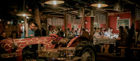 Rustic restaurant with people dining at long tables, tractor bar and hanging lamps in warm light
