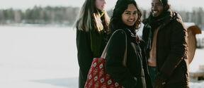 Three friends in winter coats stand smiling outdoors on snow, one with a red patterned tote bag.