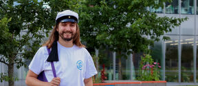 Jacek Pawlak with a student cap and purple backpack stands on green campus lawn by trees and modern building