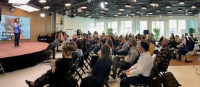 Speaker in blue jacket on stage addressing a full audience in a bright modern conference room