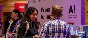 People talk at a tech fair booth, one with a backpack, another in blue checked shirt, machines beside them