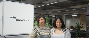 Two young women stand smiling in a modern Aalto Founder School office, with plants and desks behind them.
