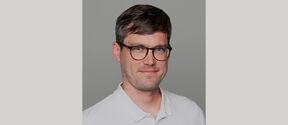 Smiling man with short brown hair and glasses in a white polo shirt, against a plain grey background