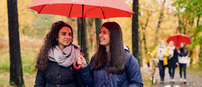 Two women share a red umbrella while walking on a tree-lined path in autumn, with others in the background.