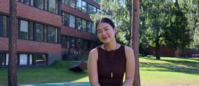 Smiling young woman on a green bench in a sunny campus park with brick buildings and tall trees.