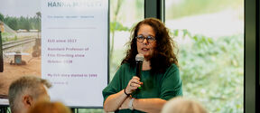 Curly-haired woman speaks into a microphone indoors, standing next to a screen.
