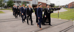 People in dark suits walking on campus across tram tracks. Väre building in the background.
