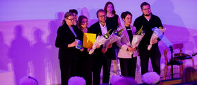 Award ceremony on purple-lit stage, group holding flowers and certificates facing audience in white caps
