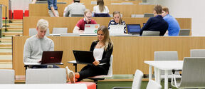 Students working on laptops in a tiered, modern classroom with light wood and grey chairs