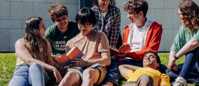 Group of students sitting on grass outside a building, reading books and chatting in the sun