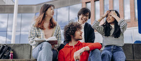 Four students sitting on outdoor steps, talking and laughing with books and a tablet by modern buildings
