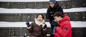 Three friends in winter coats sit on snowy steps, laughing and eating pastries with hot drinks.