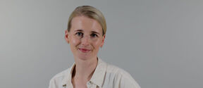 Smiling woman with light hair in a cream shirt, standing against a plain light grey background