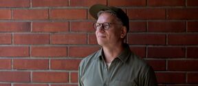 Smiling person wearing glasses, green shirt and cap, standing against a red brick wall