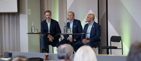 Heikki Lempinen, Jukka Salonen and Munir Mandviwalla with microphones sit at a panel table, speaking to an audience in a modern hall.