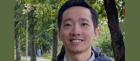 Smiling person in a jacket outdoors on a tree-lined path with green foliage in the background