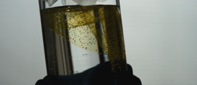 Gloved hands hold a clear beaker of yellow liquid with bubbles and a white filter paper on top