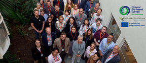 Group of conference participants with name badges, standing indoors beside plants and EU project logos