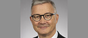 Smiling middle‑aged man in dark suit and tie, round black glasses, short grey hair, neutral grey background