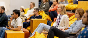 Participants in the Inventors programme sit on yellow beanbags and wooden blocks in a modern room, listening to a talk in the AGrid building at Aalto campus. 