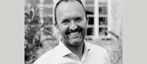 Black-and-white portrait of a smiling bearded man in a light shirt, standing outdoors before a blurred building