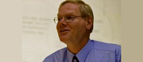 Middle‑aged man with glasses in a light blue shirt and striped tie, smiling and looking sideways indoors