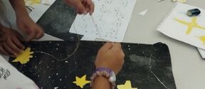 Children craft a starry night collage, gluing yellow stars onto a large black paper sky