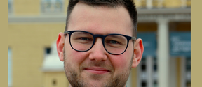 Smiling man with dark hair, beard and black glasses outdoors in front of a yellow building