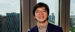 Smiling person with glasses in a blue blazer stands indoors by tall windows overlooking a city skyline.