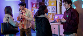 Four people talk at a tech event, two by a laptop, colourful UN goal posters glowing in the background