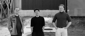Three smiling men standing outdoors in black and white, hands in pockets, industrial buildings behind