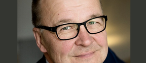 Middle‑aged man with black glasses smiling softly, close‑up indoors with neutral background