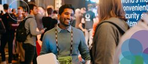 Smiling man with badge talks to a woman at a busy tech event, bright lights and screens in the background
