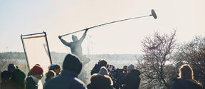 Outdoor film crew in winter clothes, boom mic raised high above a busy set near bare trees