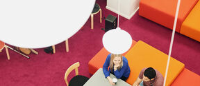 Tre personer samtalar vid ett bord i en färgstark lounge med rosa matta, orange soffor och bärbara datorer