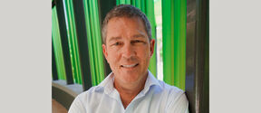 Smiling middle‑aged man in a white shirt, seated by tall green and dark columns outdoors