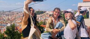 Three young people take a selfie on a sunny city viewpoint, with rooftops and trees stretching behind them.