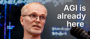 Middle‑aged man in glasses speaks into a black studio microphone, text reads “AGI is already here”.
