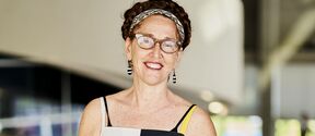 Smiling woman with glasses in patterned dress and headband, standing indoors in soft light