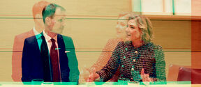 Double exposure of man in suit and woman in patterned jacket talking at a light wood meeting table